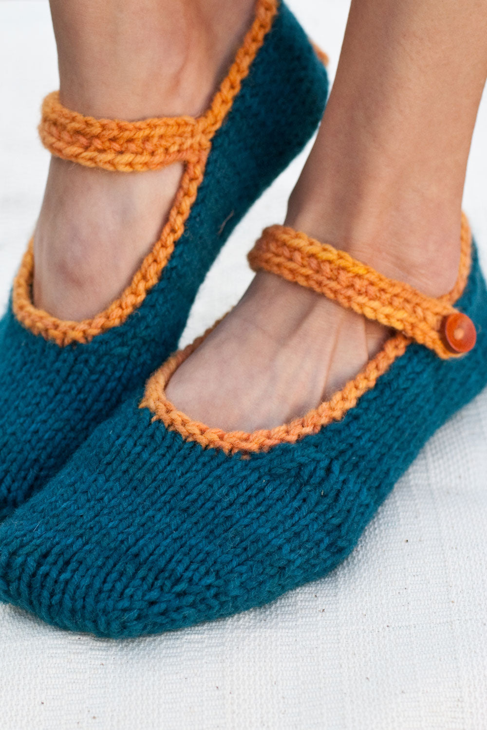 woman's feet wearing dark teal hand-knit slippers with bright orange trim, an ankle strap and wooden buttons