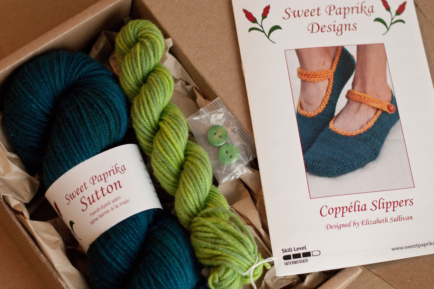 open cardboard box with kit components: pattern, large skein, mini skein and buttons